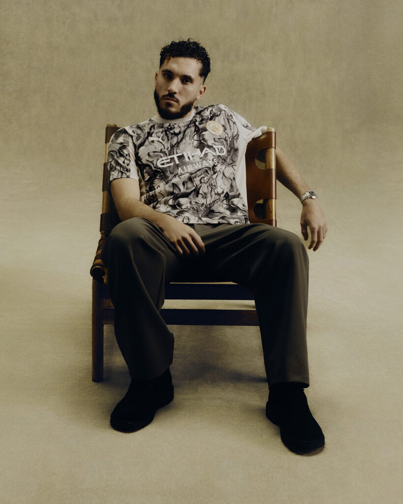 Man seated on a chair wearing a patterned shirt and dark pants in a neutral background. - Totalsports