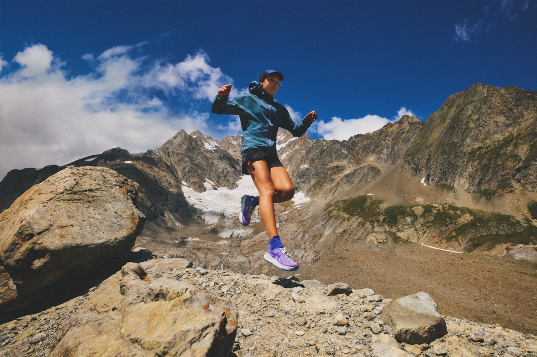 Person mid-air on a rocky alpine trail under a blue sky, wearing teal jacket, dark shorts, and purple sneakers while mountains rise in the background - Totalsports