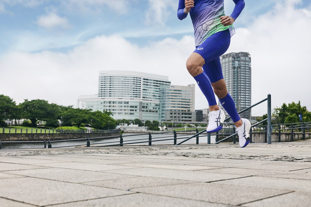 ASICS branded running scene featuring a athlete in blue tights mid-stride on a city waterfront promenade with modern buildings and a bright sky. - Totalsports