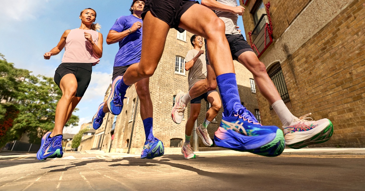 Group of runners in blue athletic sneakers sprinting along a sunlit outdoor street with brick buildings in the background, capturing motion and athletic energy. - Totalsports