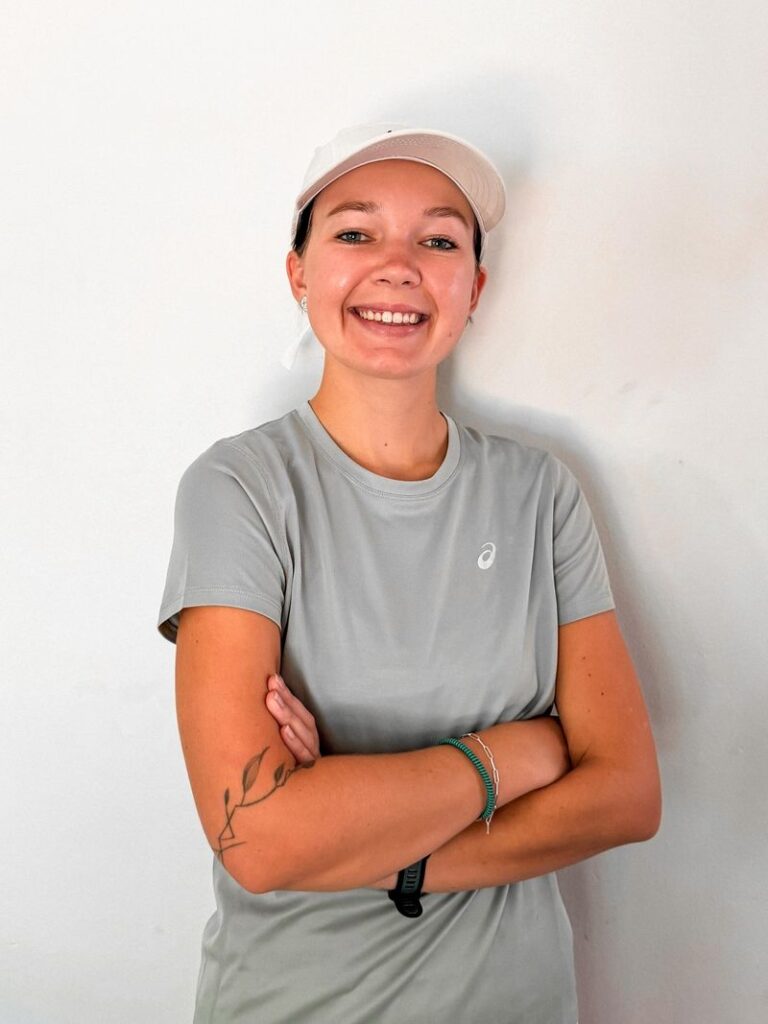 Smiling woman in a light gray athletic t-shirt and white cap stands against a plain white wall, arms crossed, with a small tattoo visible on her left forearm. - Totalsports