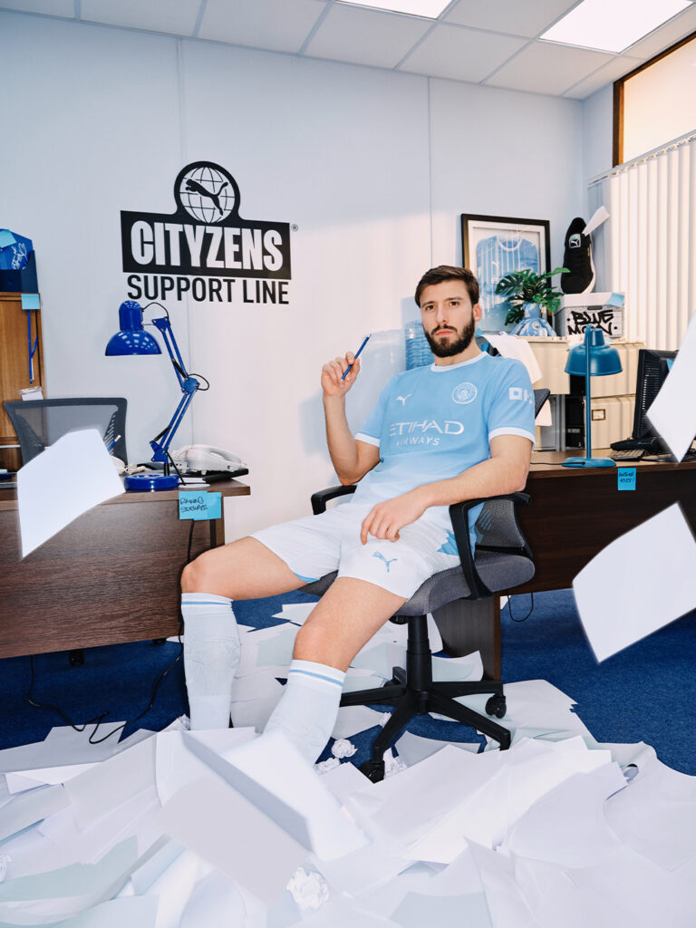 Man City home matchwear in light blue and white is worn by Dias in an office setting beside a Citizens Support Line sign and scattered papers. - Totalsports