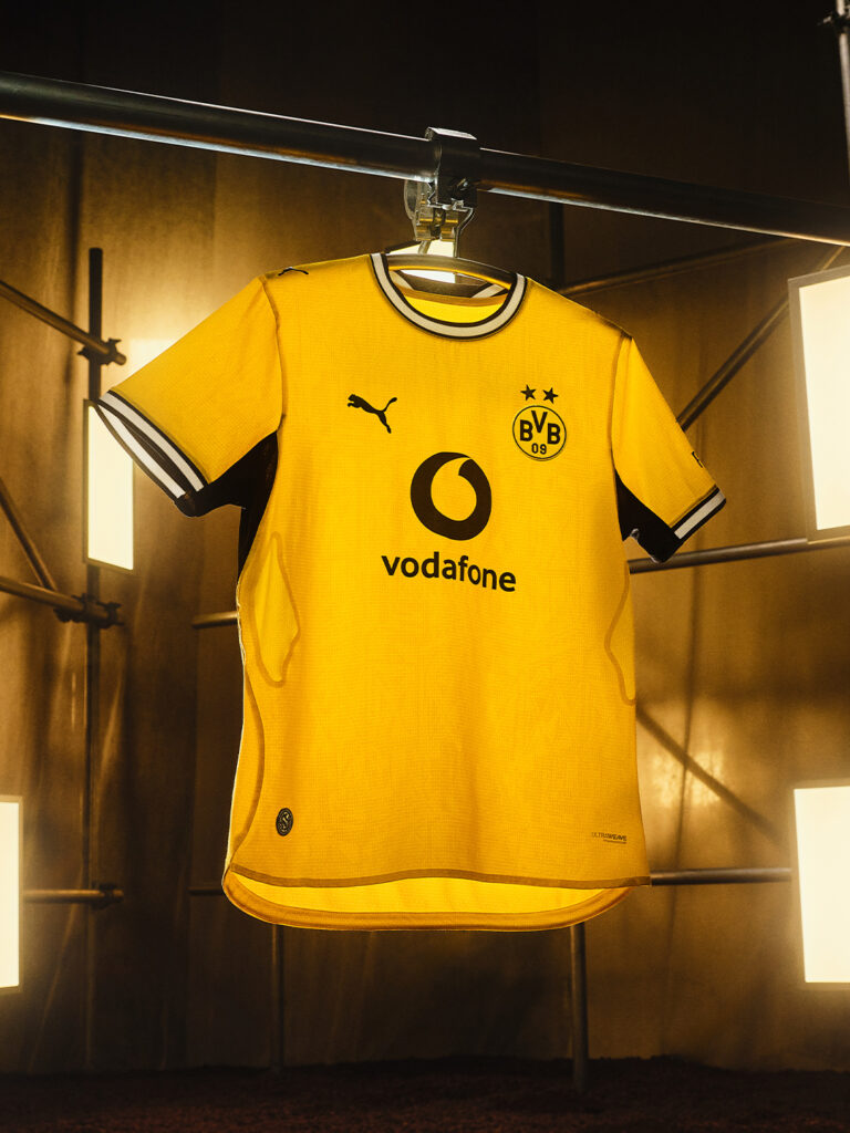 Yellow BVB home matchwear football jersey with black Puma details and sponsor print, hanging under warm studio lights - Totalsports