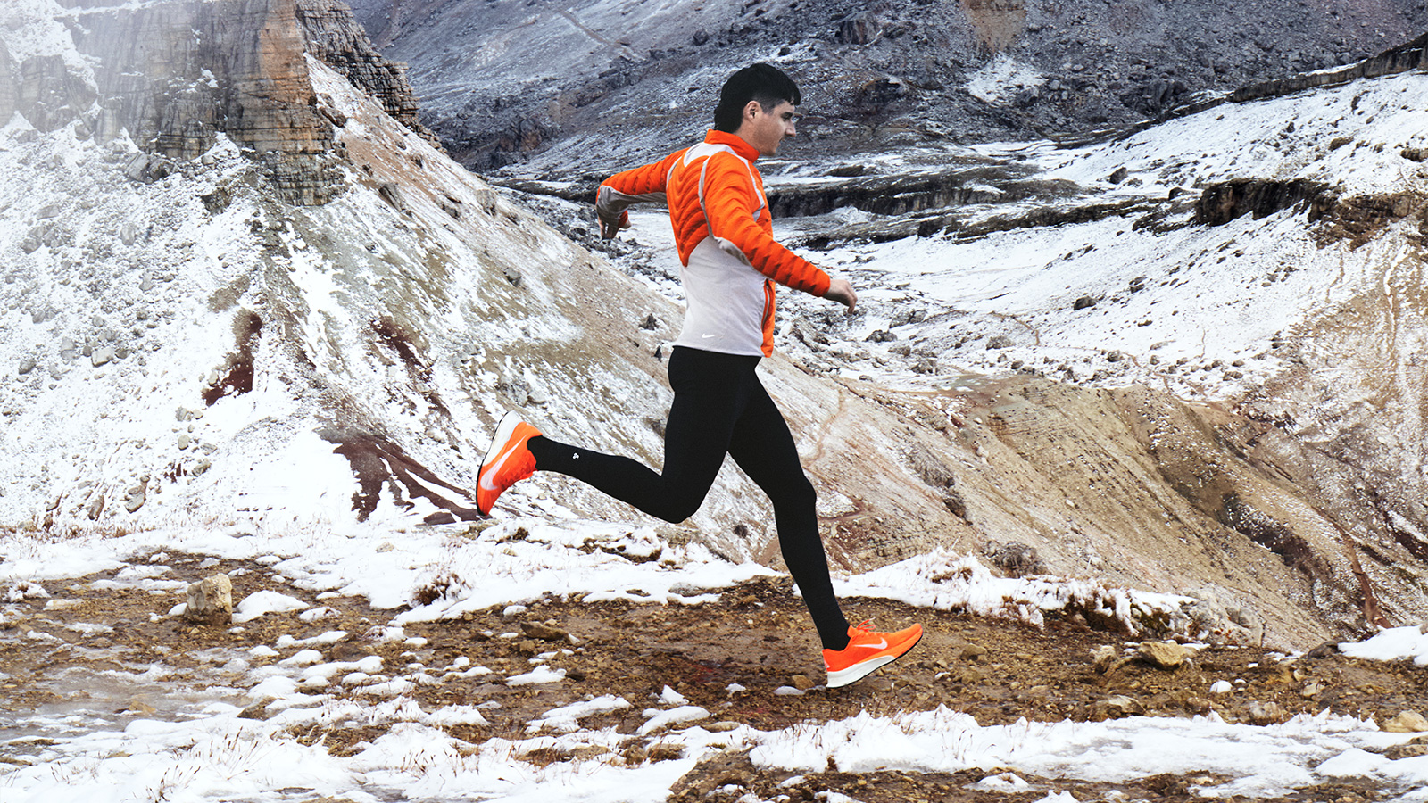 A runner in orange gear strides across snowy rocky terrain wearing Nike Ultrafly 2 running shoes - Totalsports