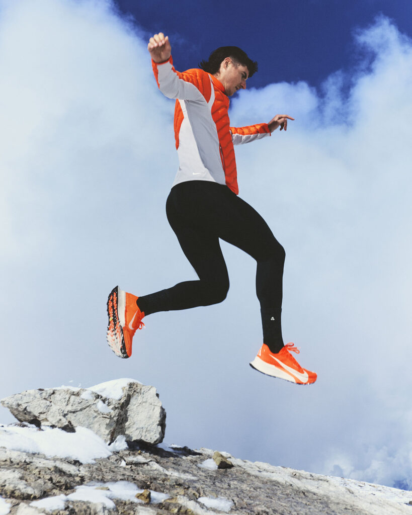 Running jump in snowy conditions with Nike Ultrafly 2 trainers, orange jacket and black tights against a cloudy sky - Totalsports