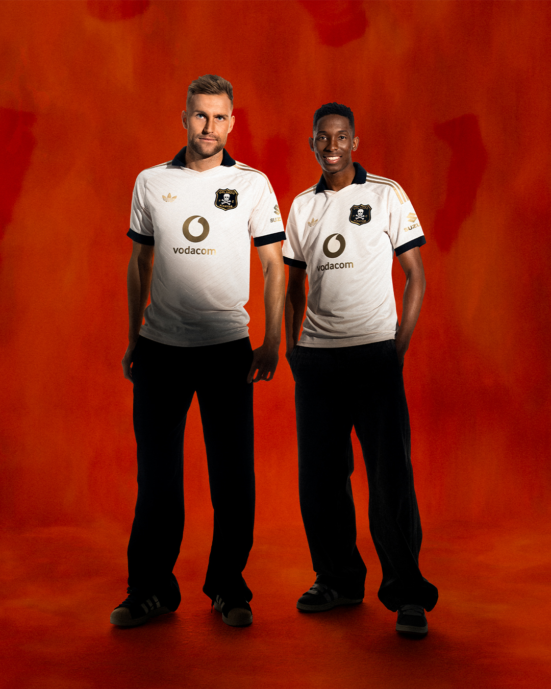 Two men stand side by side in Orlando Pirates home jerseys with Vodacom branding against a vivid red backdrop. - Totalsports